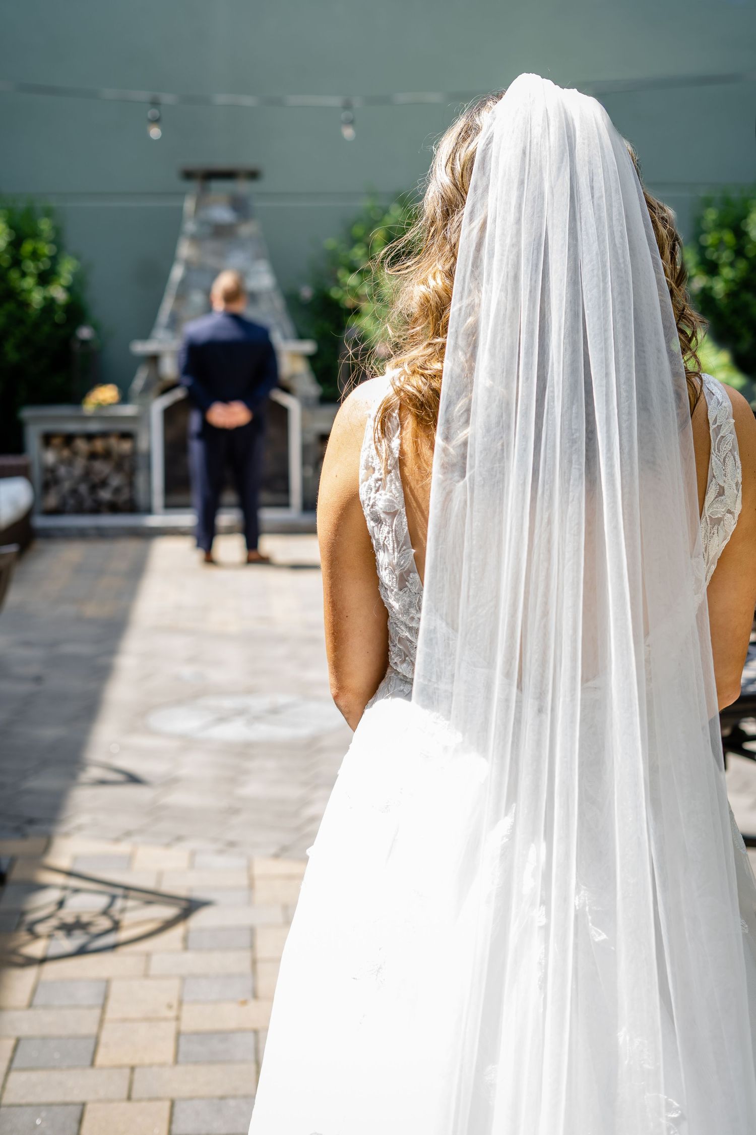 Back view of bride in white wedding dress and long veil looking towards officiant during outdoor ceremony.