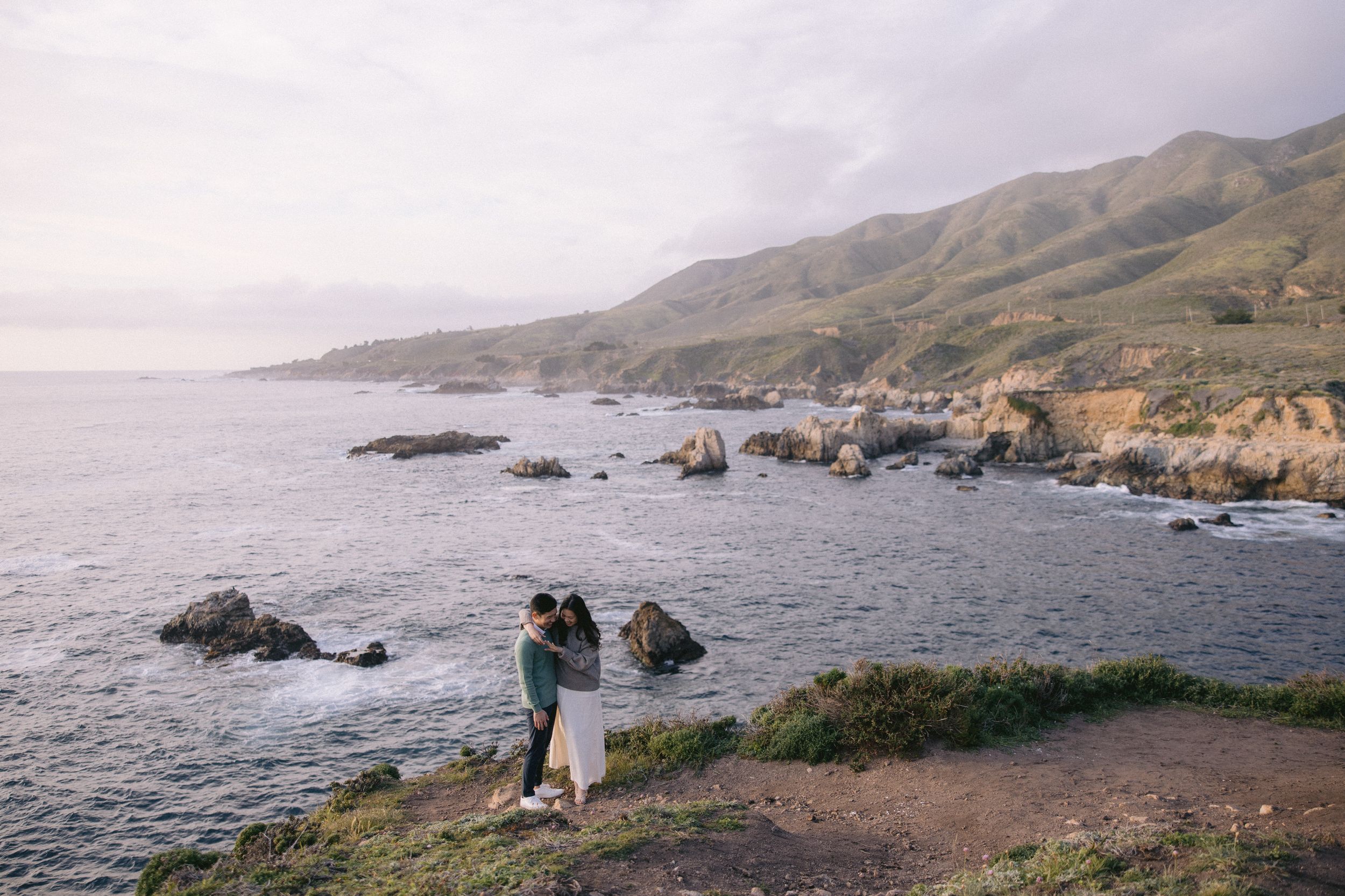 Couple admiring the rugged Big Sur coastline with rocky cliffs and crashing waves along California's Pacific Coast Highway.