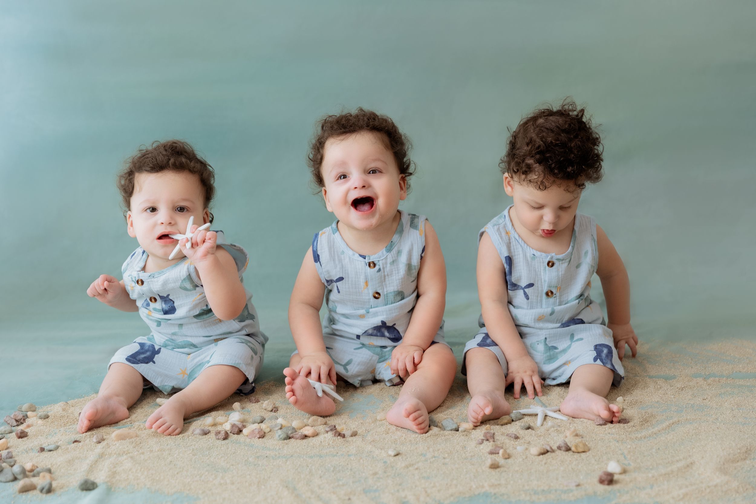 Three babies wearing matching blue rompers sit on sand against a turquoise backdrop showing different expressions.