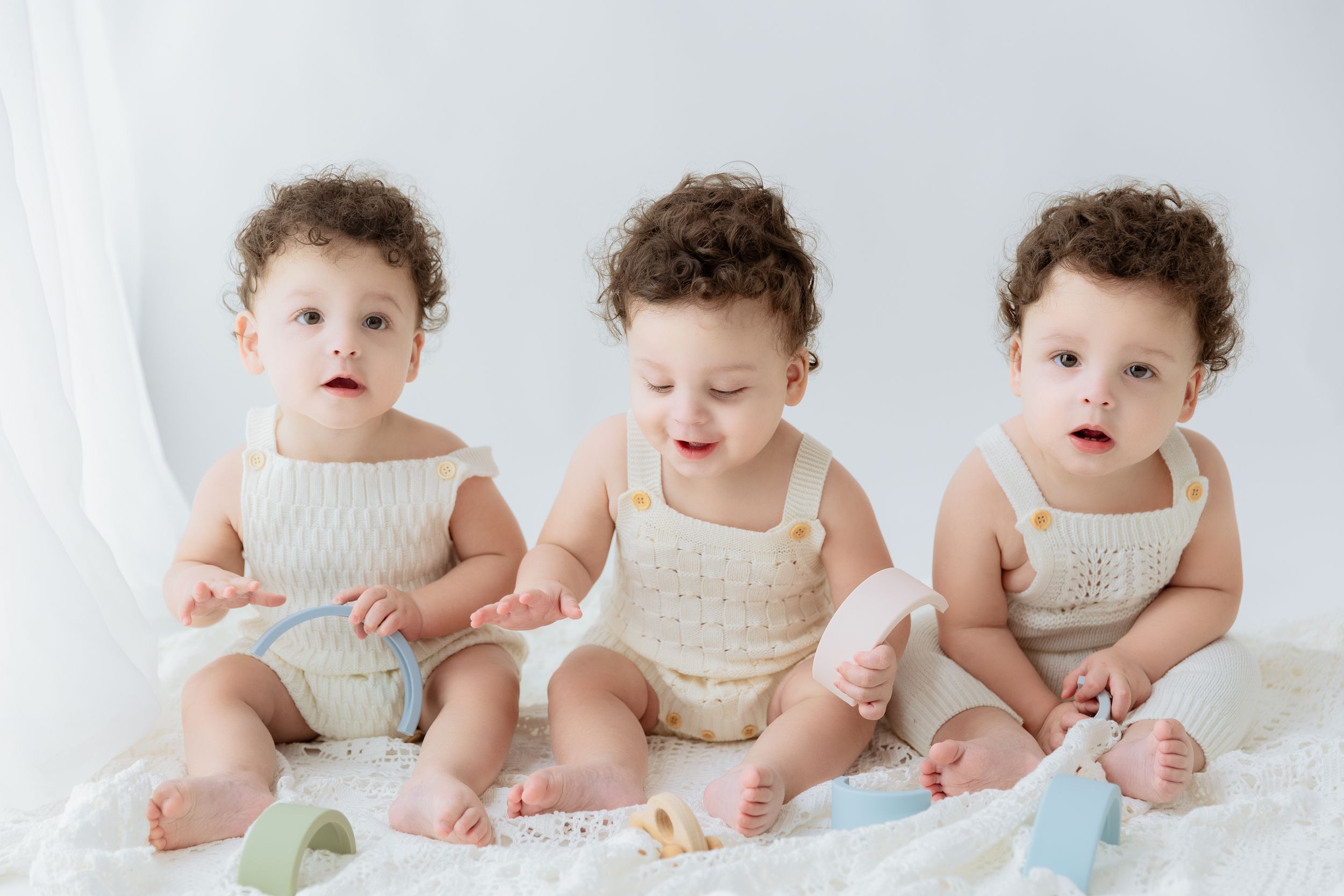 Three babies wearing matching cream-colored outfits sit together on white fabric while posing for a portrait.