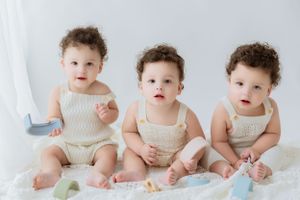 Three babies in white knit rompers sitting on white fabric with soft toys.