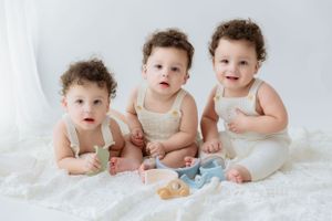 Three infants in cream-colored outfits playing with pastel toys on white blanket.