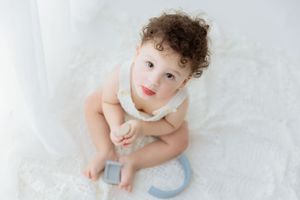 Baby in white outfit sits on soft white blanket playing with a gray and blue teething toy.