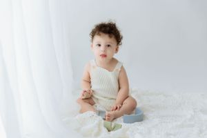 Baby wearing knit romper sitting alone with toys on white background.