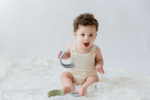 Infant wearing cream-colored romper sits on white fluffy rug looking excited and playful.