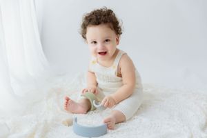 A smiling baby in a white outfit sits on white fabric with a blue bowl.