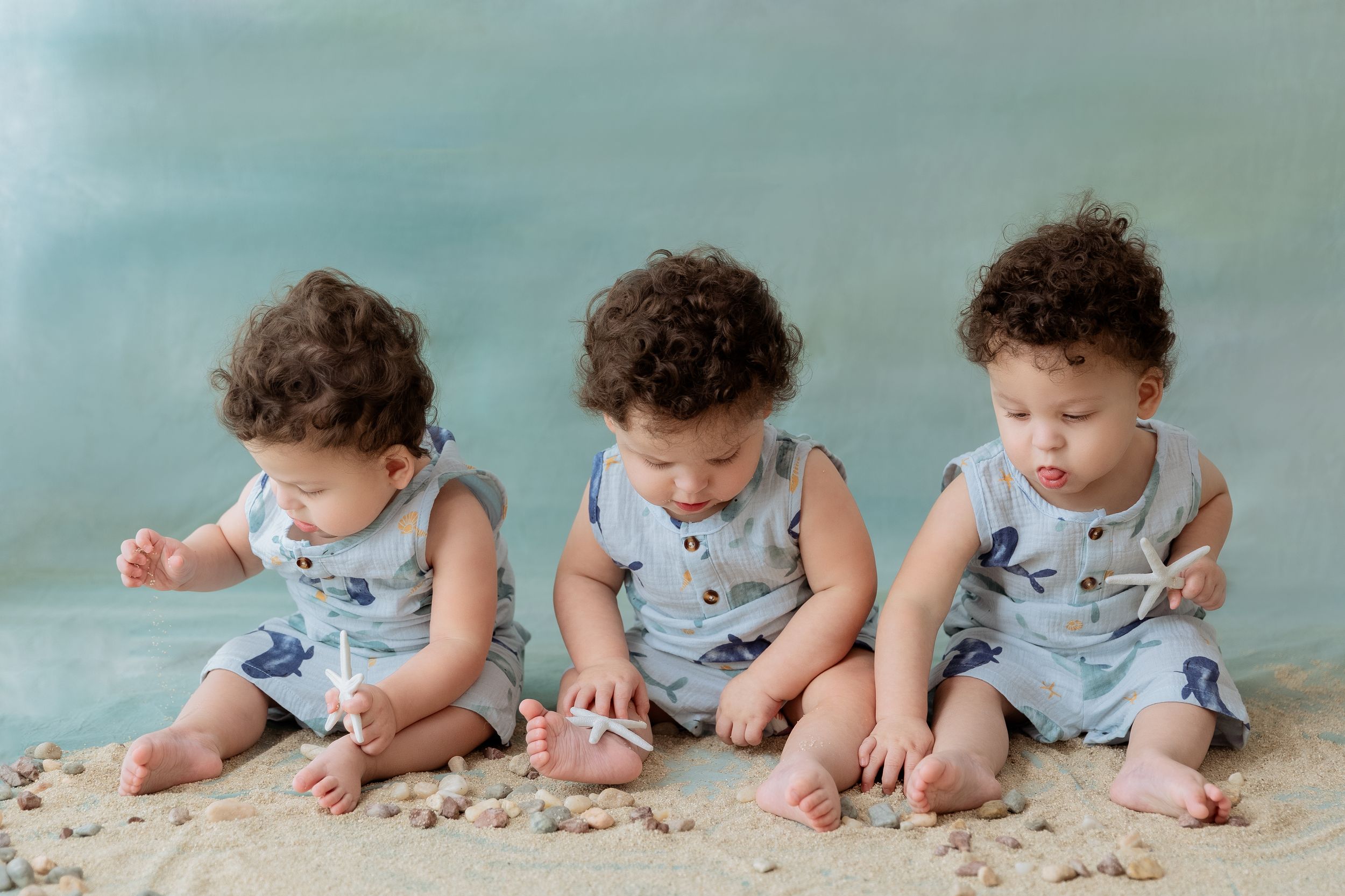 Three babies in matching blue and white overalls sit together on sand while exploring with their hands.