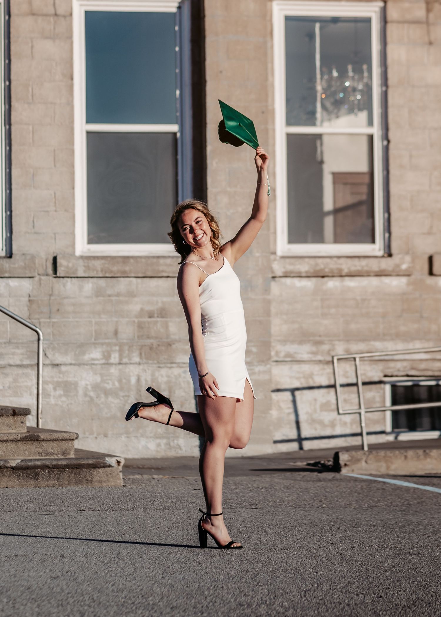 Graduate in white dress celebrates with green cap against stone building exterior on sunny day.