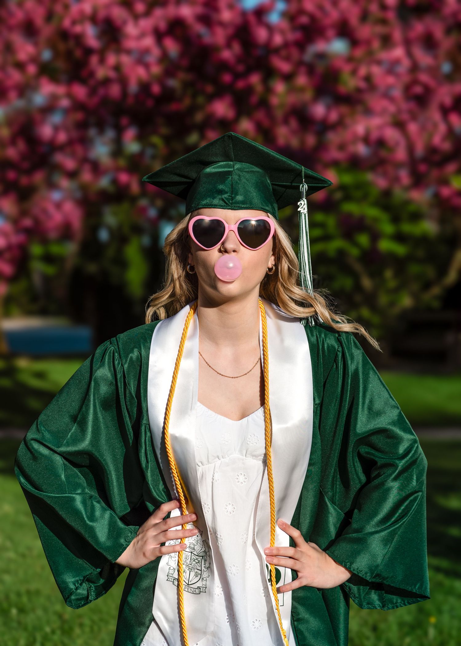 Graduation photo showing someone in emerald green cap and gown wearing heart sunglasses and making silly face with cherry blossoms behind.