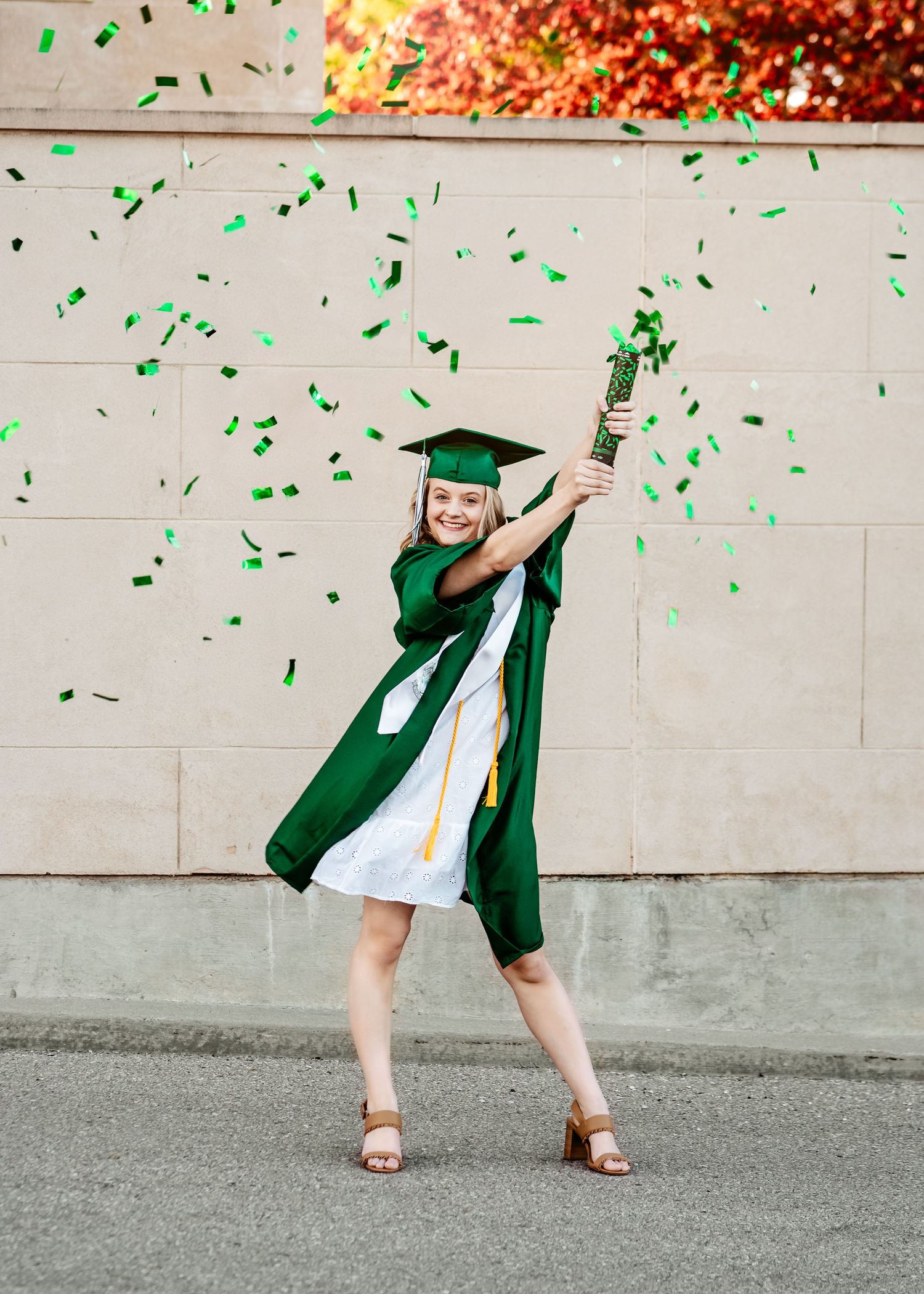 Green graduation cap and gown worn by graduate celebrating with confetti flying in joyful pose against wall.