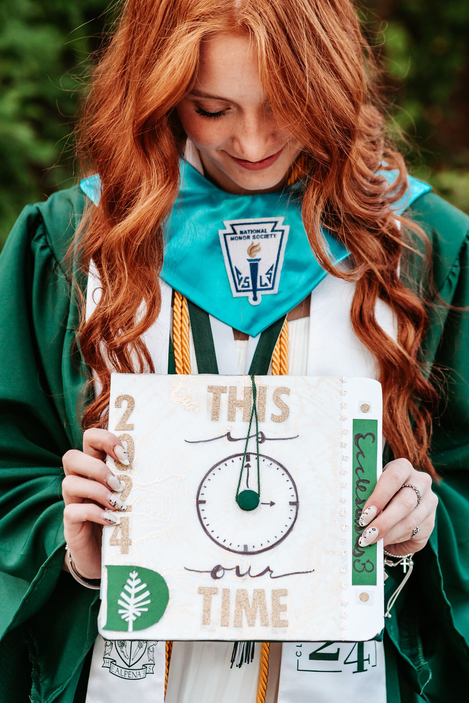 Person in green graduation gown holding a decorative clock sign with 'This Time' written on it.