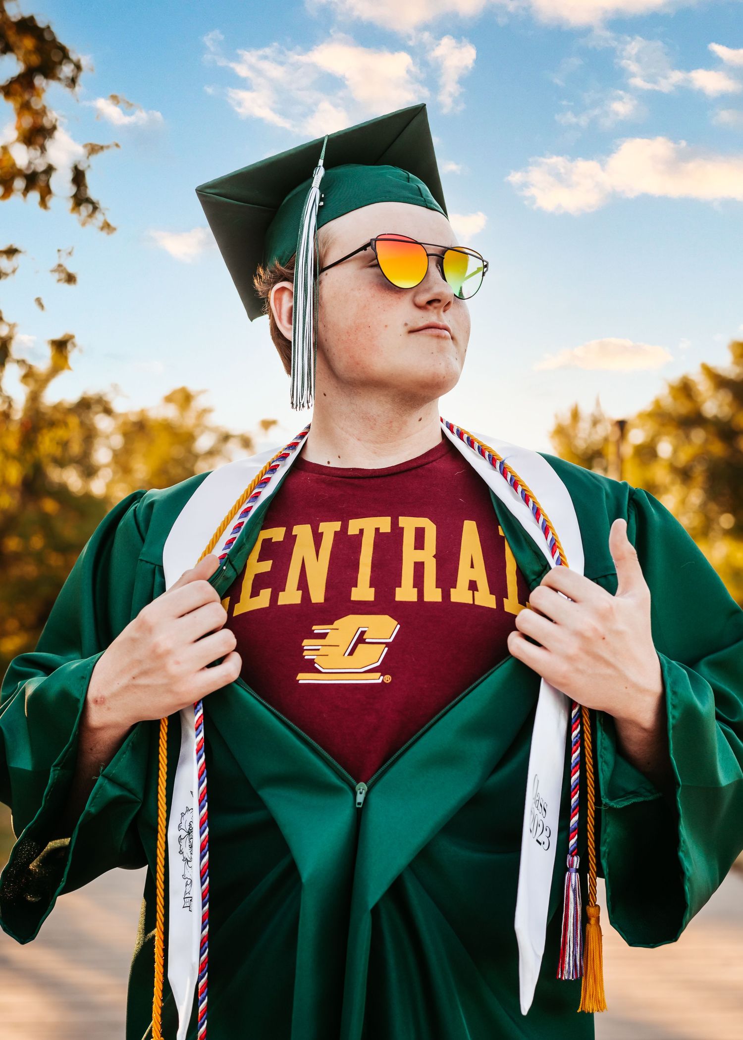 Graduate in emerald green cap and gown reveals Central university t-shirt during outdoor photo session at sunset.