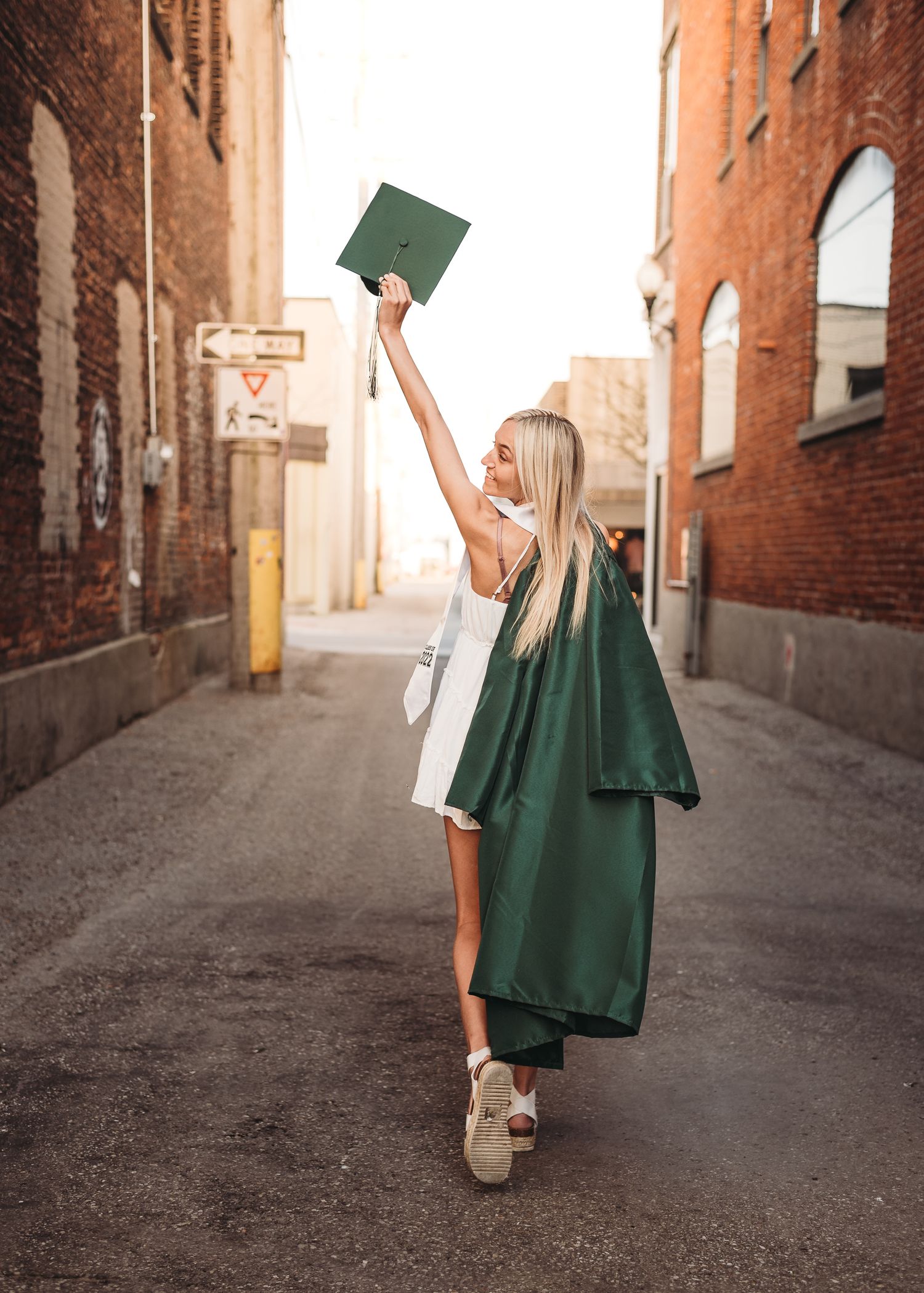 Graduate in green cap and gown celebrating with raised diploma in alleyway between brick buildings.