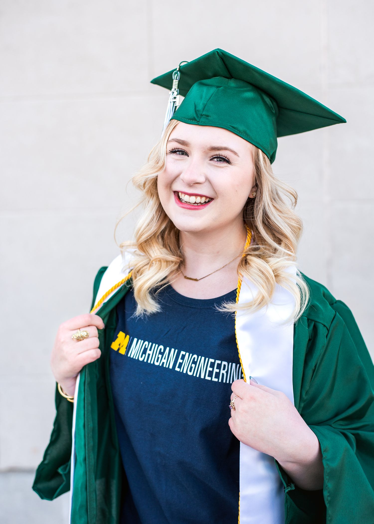 Graduate wearing emerald green cap and gown with Michigan Engineering t-shirt smiles joyfully against neutral wall.