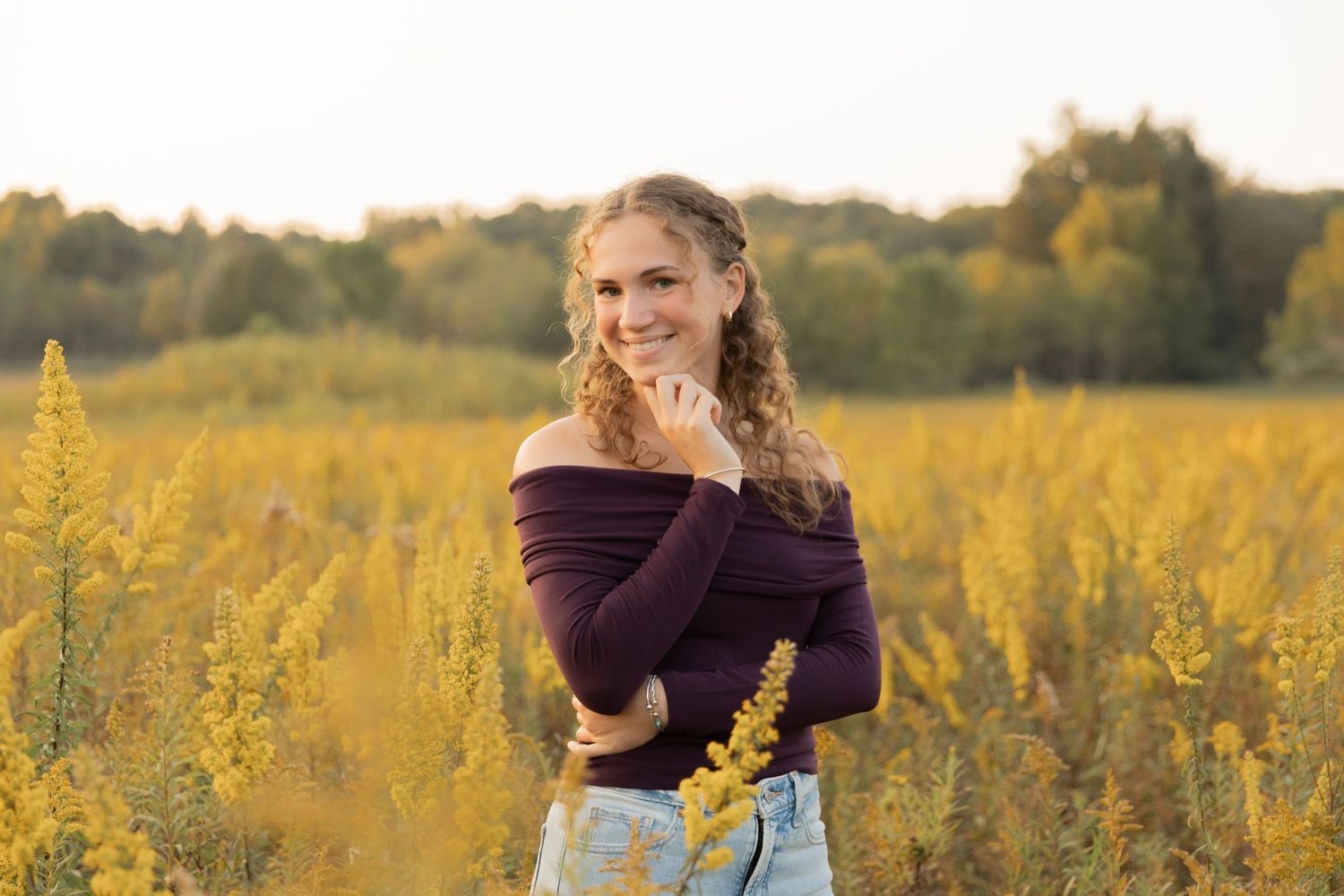 teenager in an off-shoulder sweater poses in a golden meadow at sunset with wildflowers in the background at a summer photoshoot location