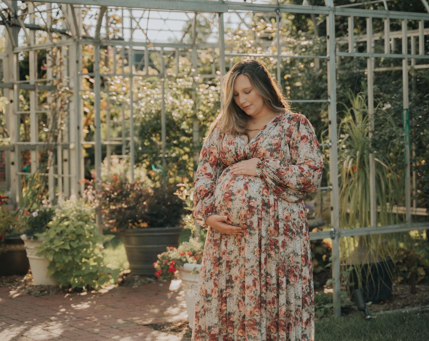 Expectant mother in a floral dress stands in a sunlit garden greenhouse.