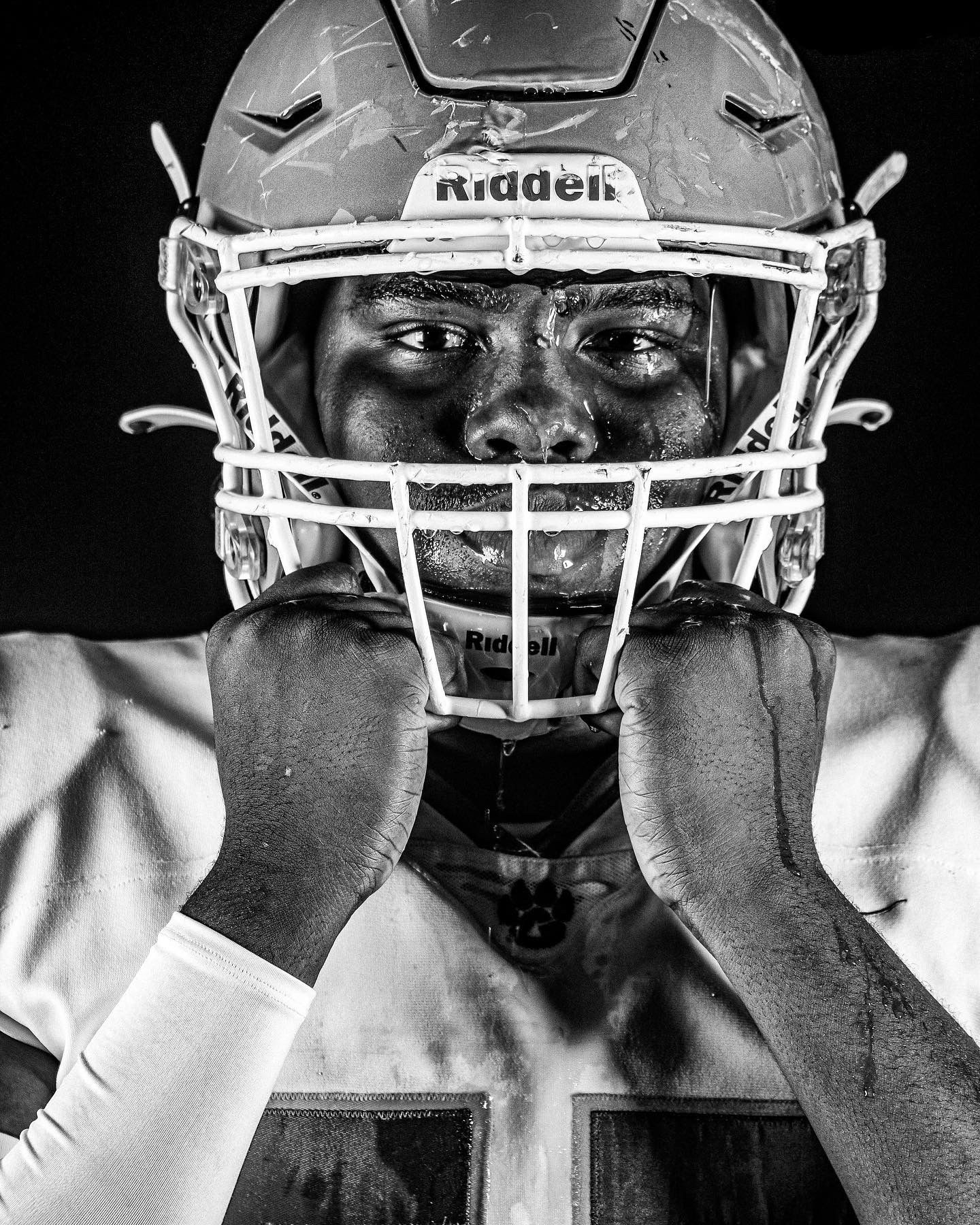 Intense black and white portrait of a football player in Riddell helmet with hands on face mask.