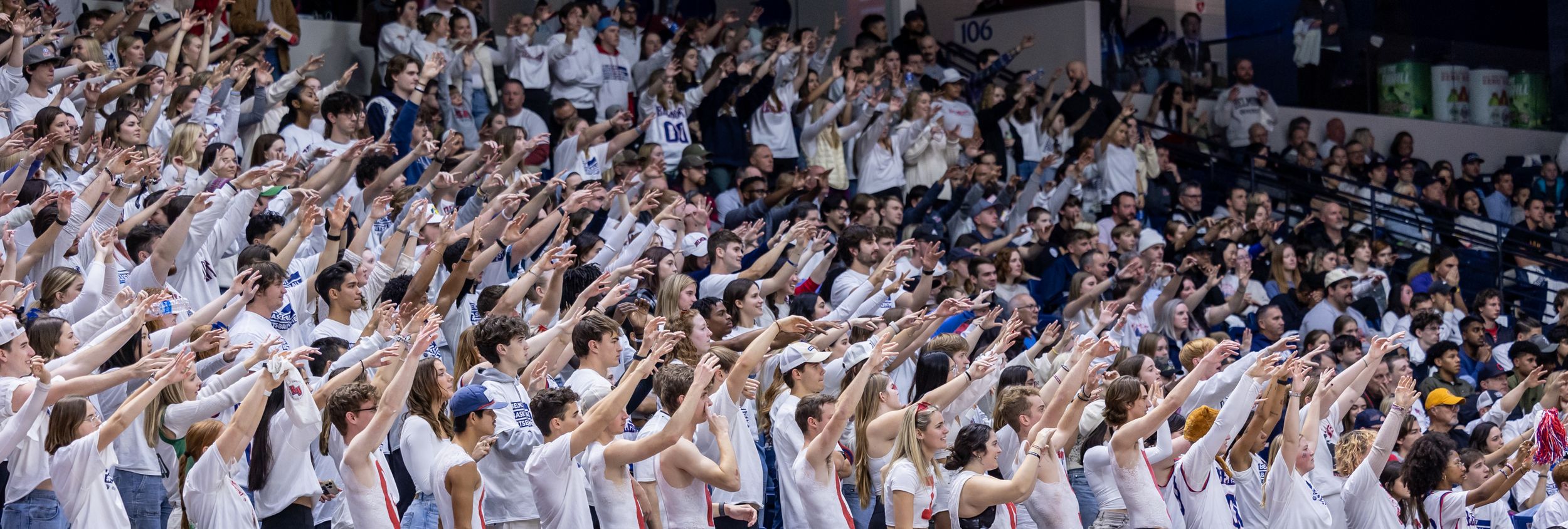 Large crowd of fans dressed in white cheering with raised arms at an indoor sports arena.