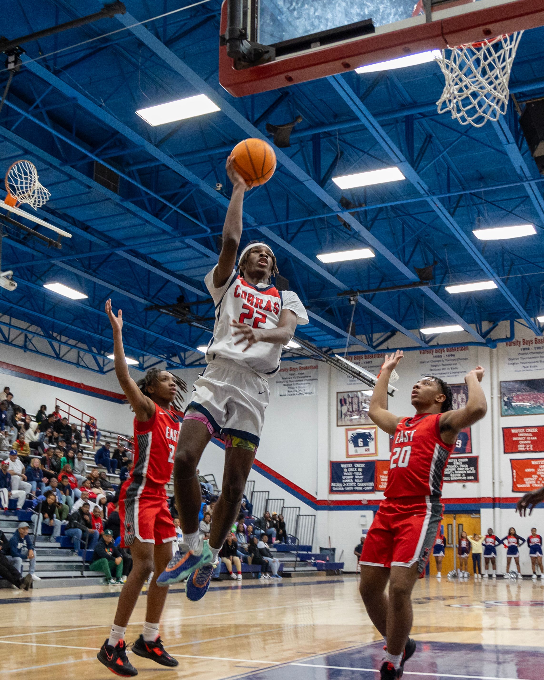A basketball player in white leaps for a layup while two defenders try to block.