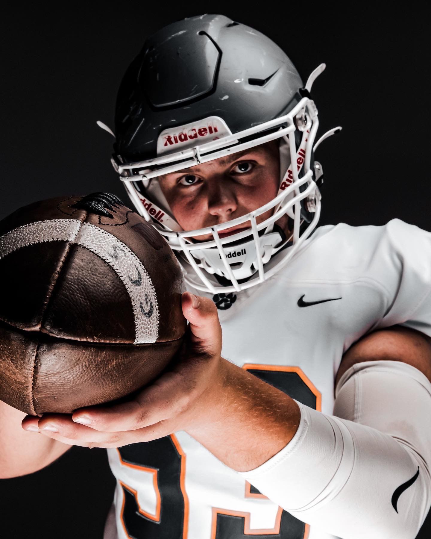 Football player in white and orange uniform holds a football toward the camera wearing a black helmet.