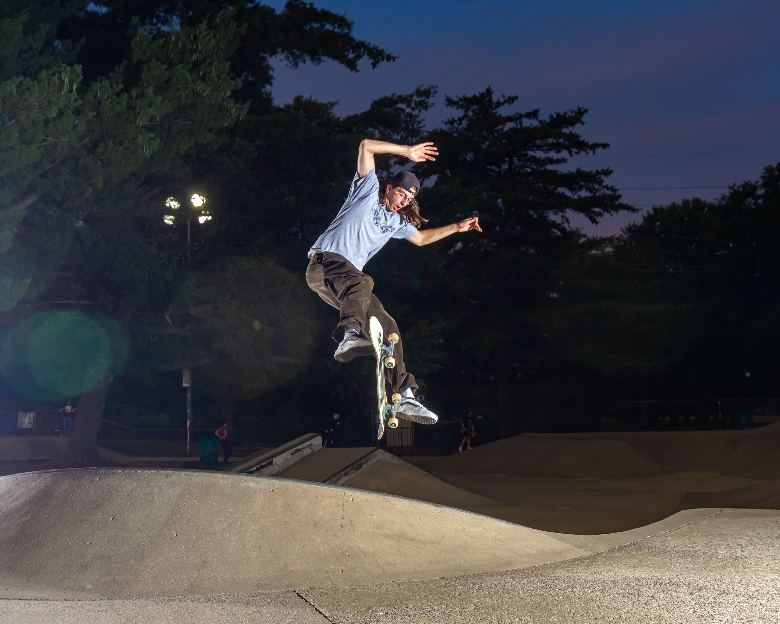 Skateboarder performs a trick at an outdoor skate park during evening with lights illuminating the scene.