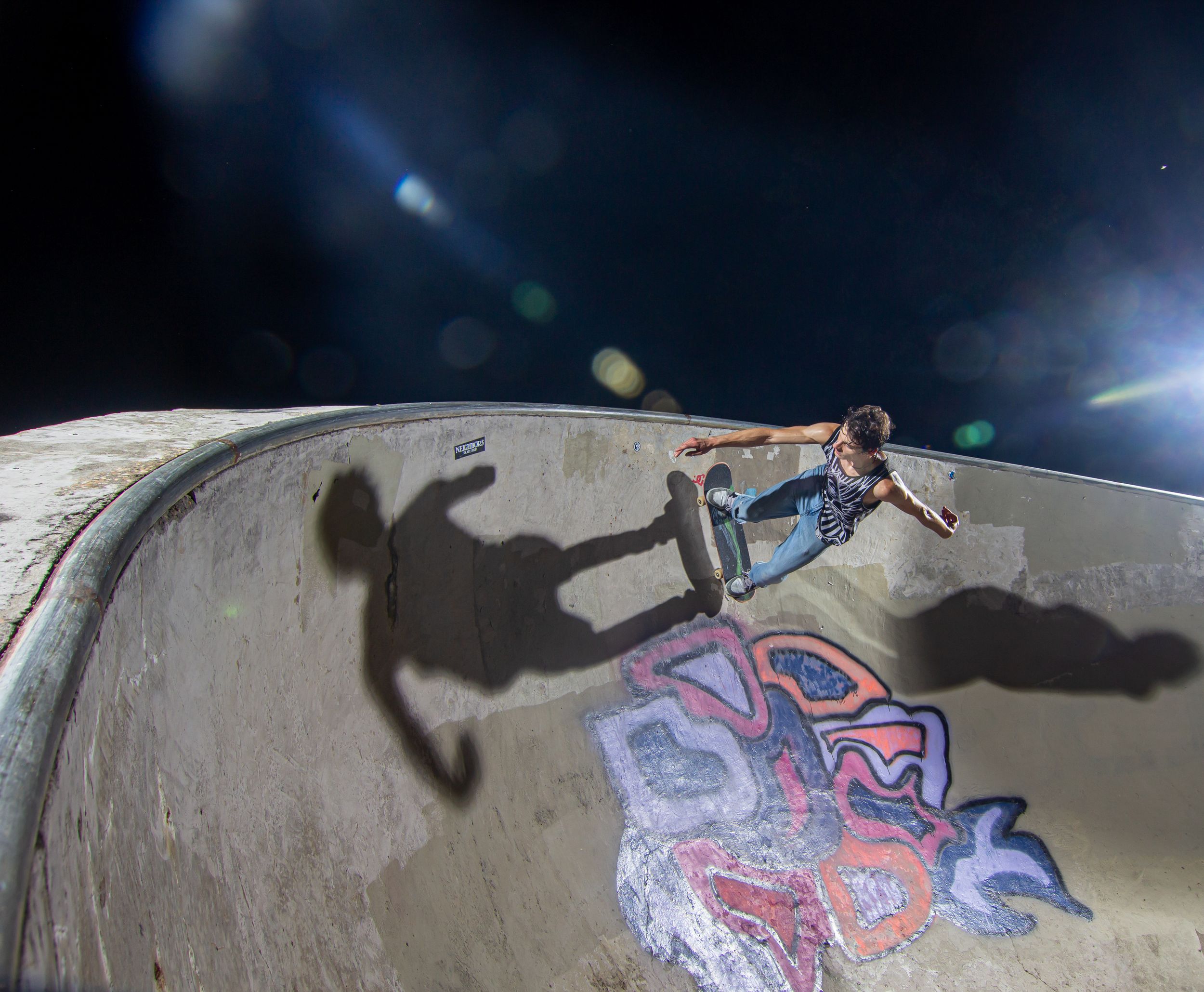 Skateboarder rides the curved wall of a skate park bowl at night with dramatic lighting and graffiti.