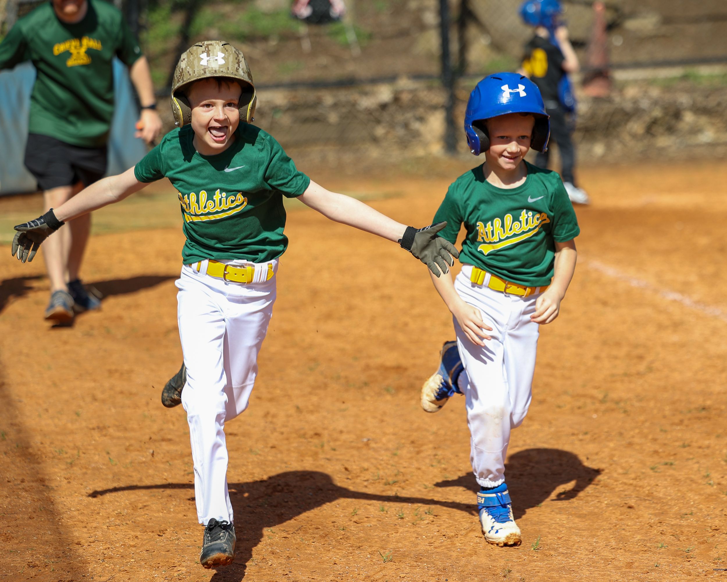 Two young baseball players in green Athletics uniforms celebrate joyfully while running the bases.