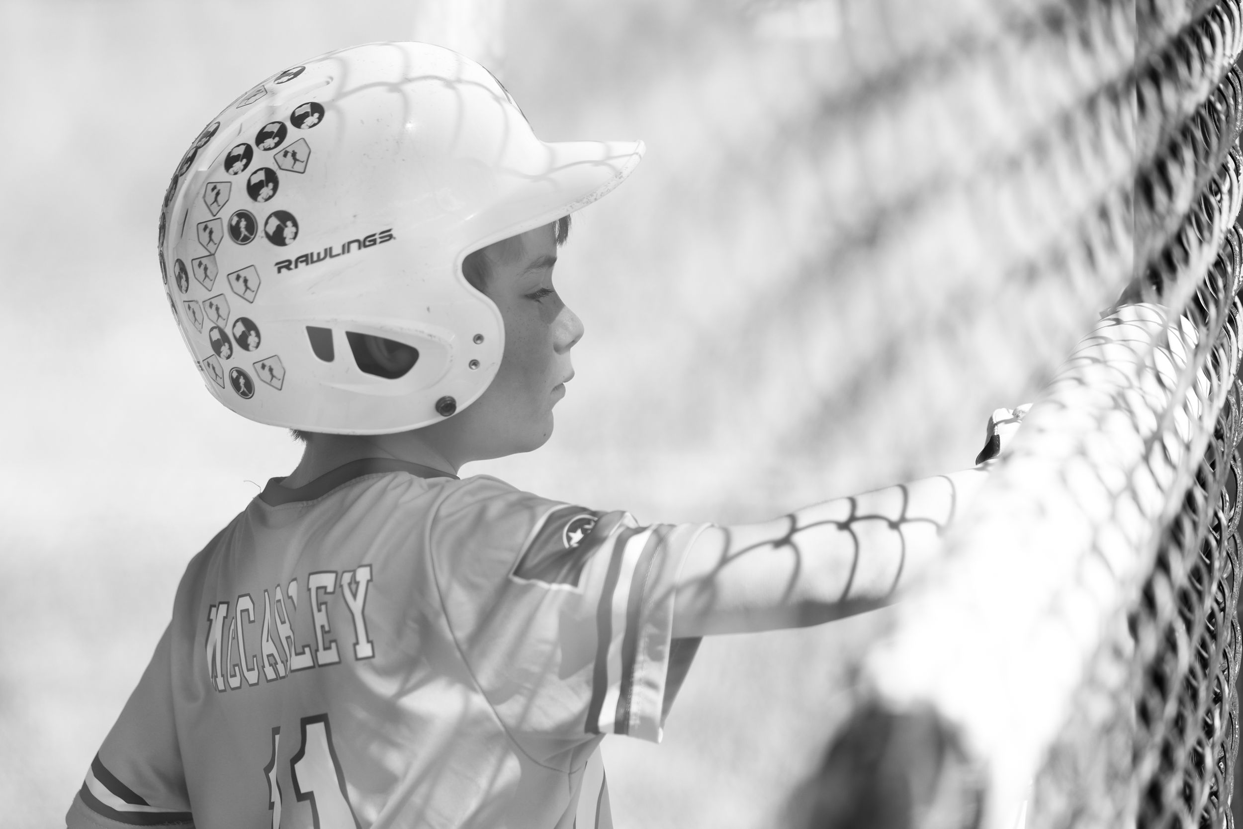 Black and white photo of young baseball player in batting helmet leaning against dugout fence.