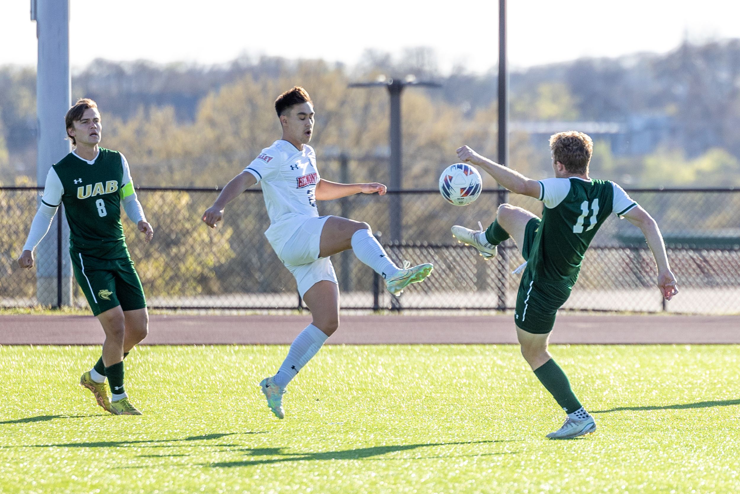 Soccer player in white uniform controls the ball between two UAB defenders during outdoor match.