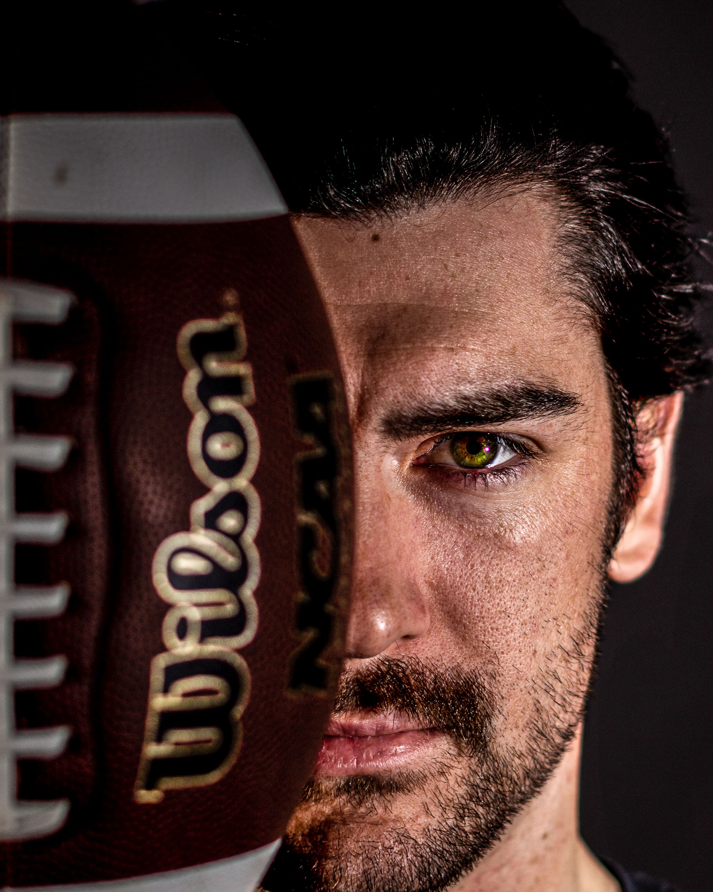 Close-up portrait of a man holding a Wilson NCAA football over half his face against a dark background.