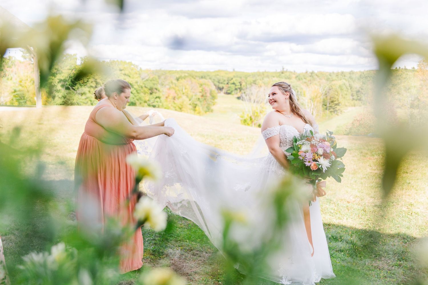 A bride in a flowing white dress shares a joyful moment in a sunlit field with someone in a coral dress.