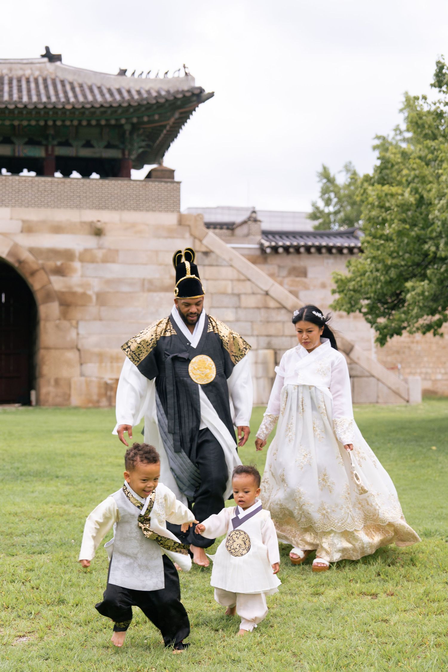 Family wearing traditional Korean hanbok attire plays together on grass near traditional palace architecture.
