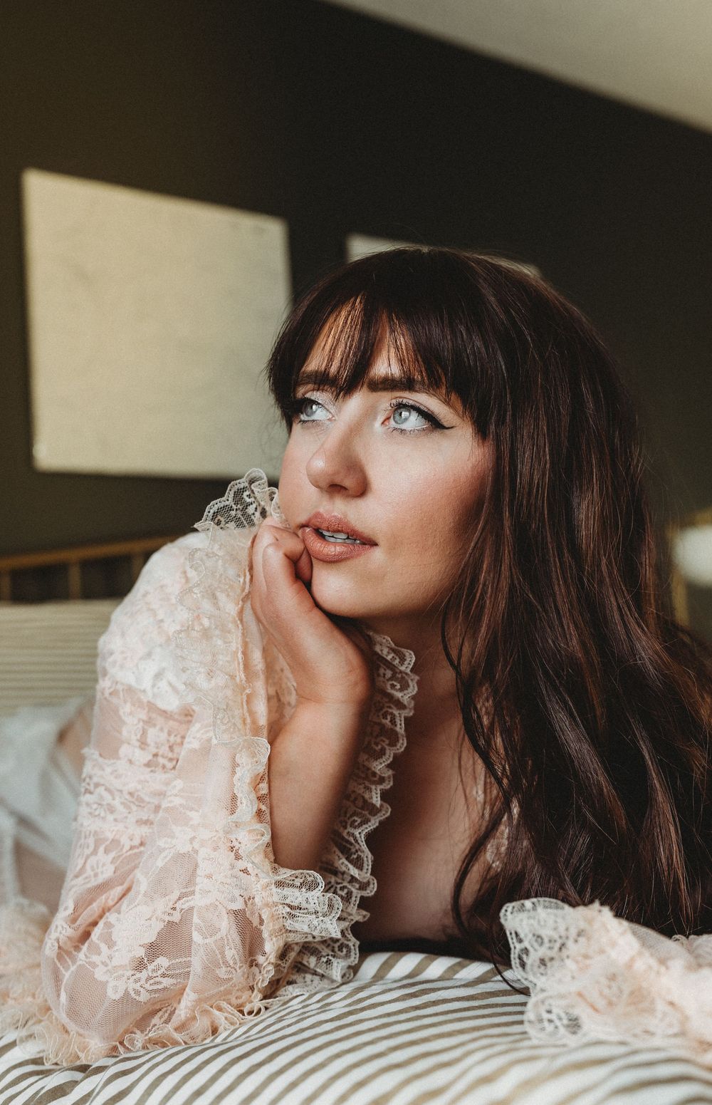 A person with dark bangs and long hair rests on striped bedding wearing a delicate lace sleeve.