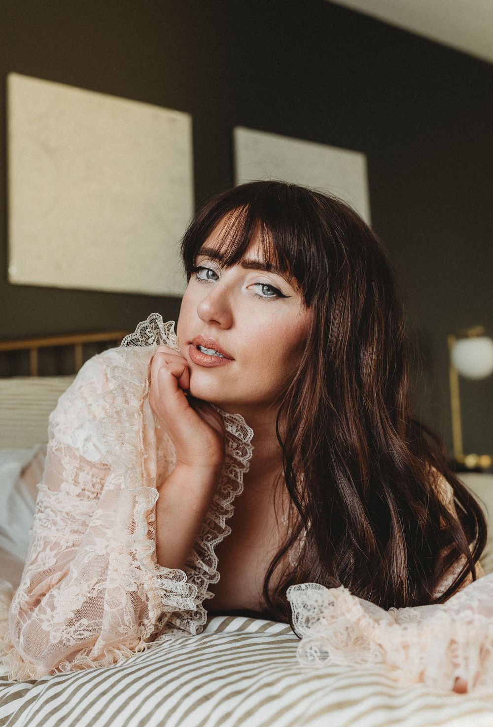 A person in a white lace dress lying on striped bedding while posing for a series of romantic boudoir photos.