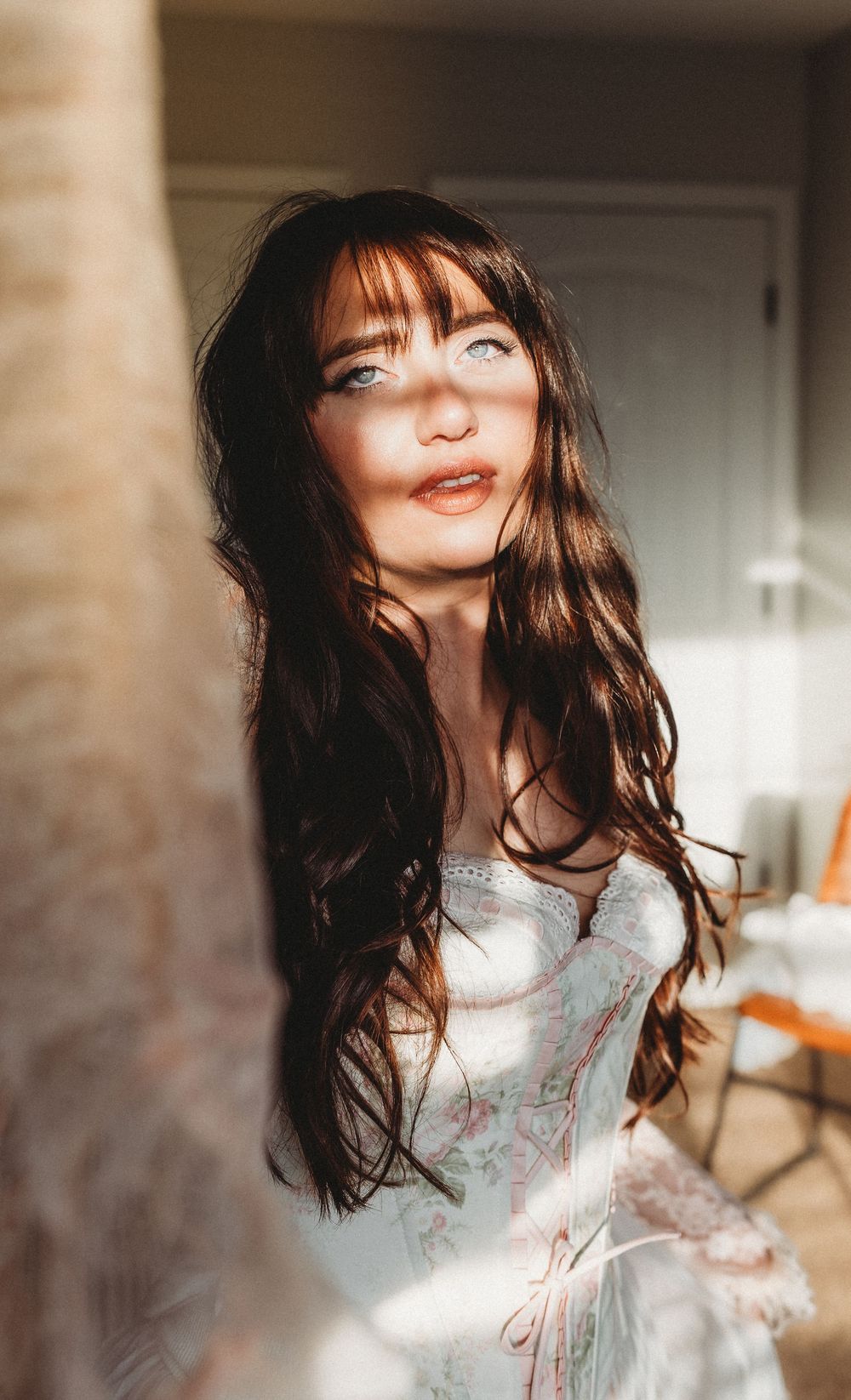 A person in a white lace dress smiles with long flowing dark hair in a warm indoor setting.