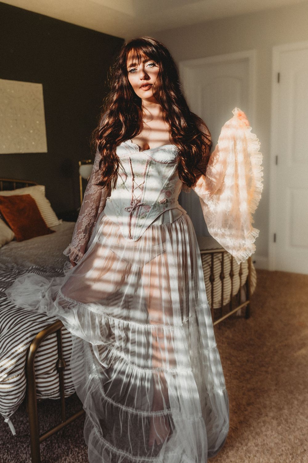 A flowing white dress creates ethereal movement in a sunlit bedroom with striped bedding and warm lighting.