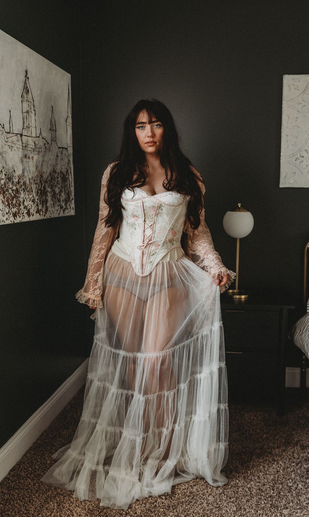 A romantic vintage-inspired gown with white corset bodice, sheer tulle skirt and long lace sleeves in a moody dark setting.