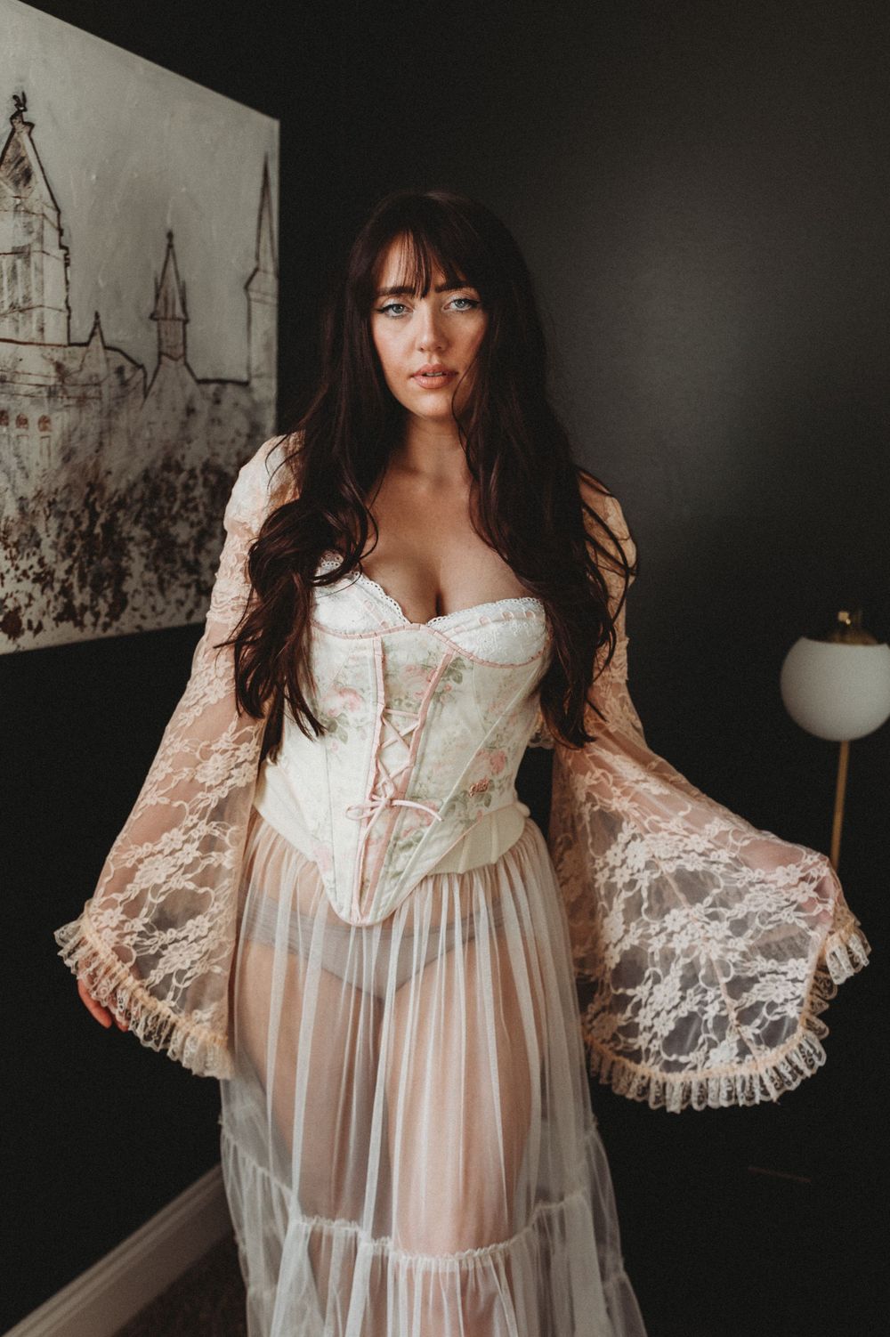 A romantic vintage-inspired white lace corset dress with sheer bell sleeves and flowing skirt stands against a dark wall.