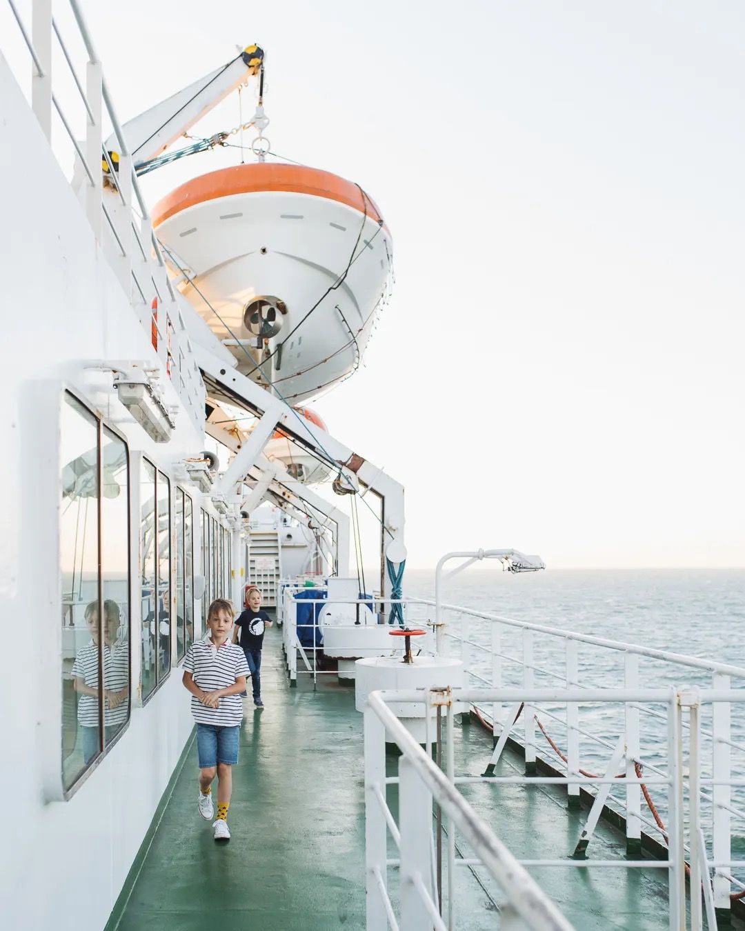 Passengers walking on a sunlit ferry deck with open sea beside them.