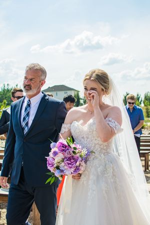 A wedding ceremony on a sunny day with purple floral bouquets and guests gathered on a wooden deck.