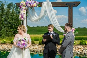 An outdoor wedding ceremony under a wooden arch decorated with white fabric and purple flowers beside a pond.