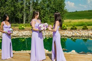 Bridesmaids in matching lavender dresses stand on wooden dock by turquoise water holding bouquets.