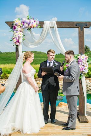 Wedding ceremony taking place under a wooden arch decorated with white fabric and floral arrangements.