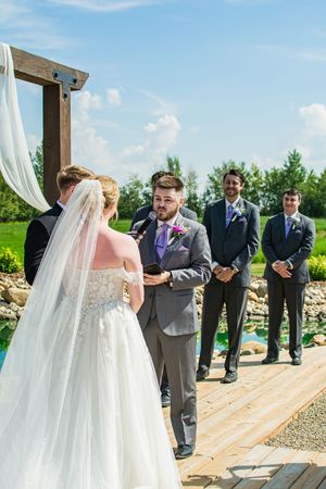 An outdoor wedding ceremony takes place under a wooden arch with flowing white fabric on a sunny day.