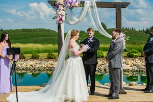 A wedding ceremony takes place at a scenic outdoor venue with a wooden arch and white draping overlooking a blue pond.