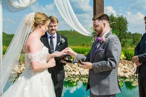 Wedding ceremony exchange of rings under white draped arch by water.