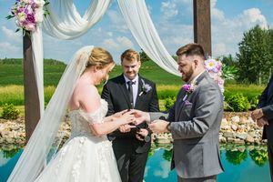 A wedding ceremony exchange of rings under a draped pergola with floral decorations and blue sky.