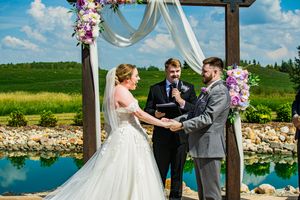 Outdoor wedding ceremony under a wooden arch decorated with purple flowers beside a serene lake.