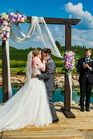 An intimate wedding ceremony moment captured under a wooden pergola decorated with white fabric and purple flowers.