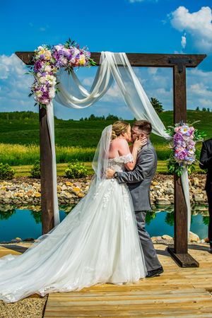 Couple shares romantic moment under wooden arch decorated with flowers and white fabric by waterfront.